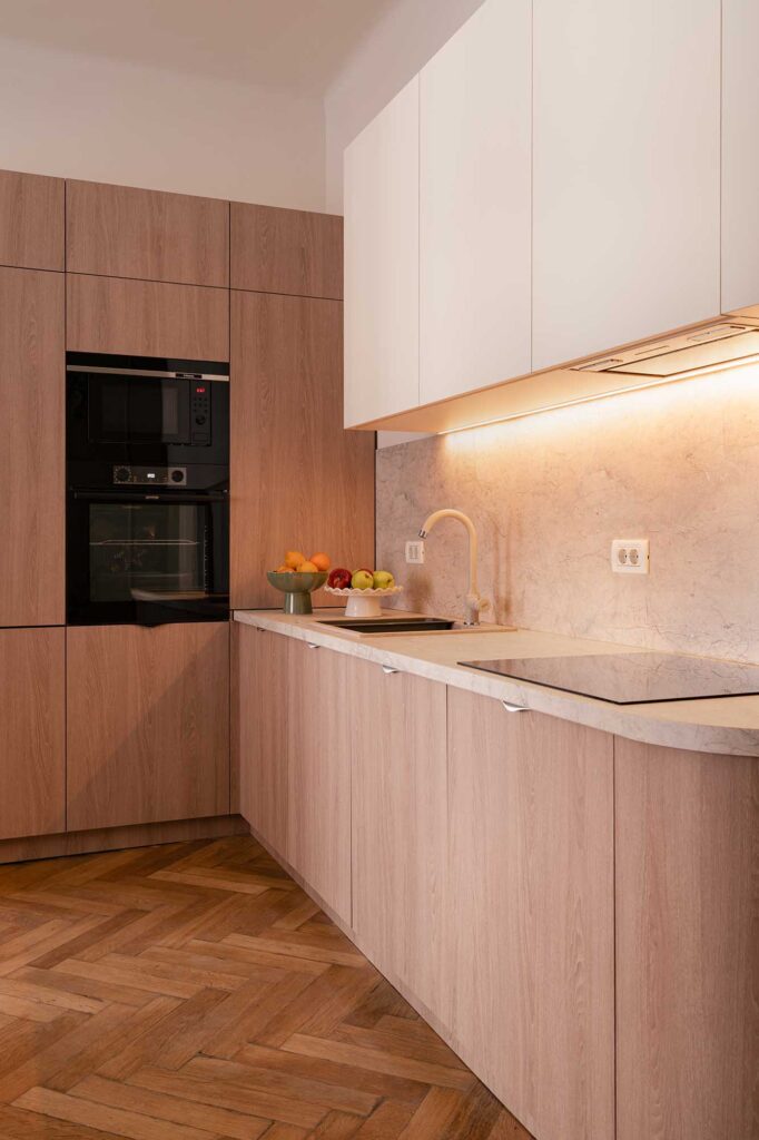 Ilisie Sergiu capture a modern curved kitchen with light wood cabinetry, white upper units, built‑in black ovens, featuring warm under‑cabinet lighting, minimalist faucet, and herringbone parquet flooring for a refined contemporary look.