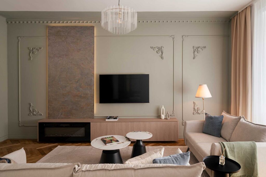 "Elegant modern living room with beige sofas, marble coffee tables, and ornate wall moldings featuring a textured stone panel and backlit TV unit, complemented by designer chandelier, warm floor lamp, and natural light through sheer curtains.