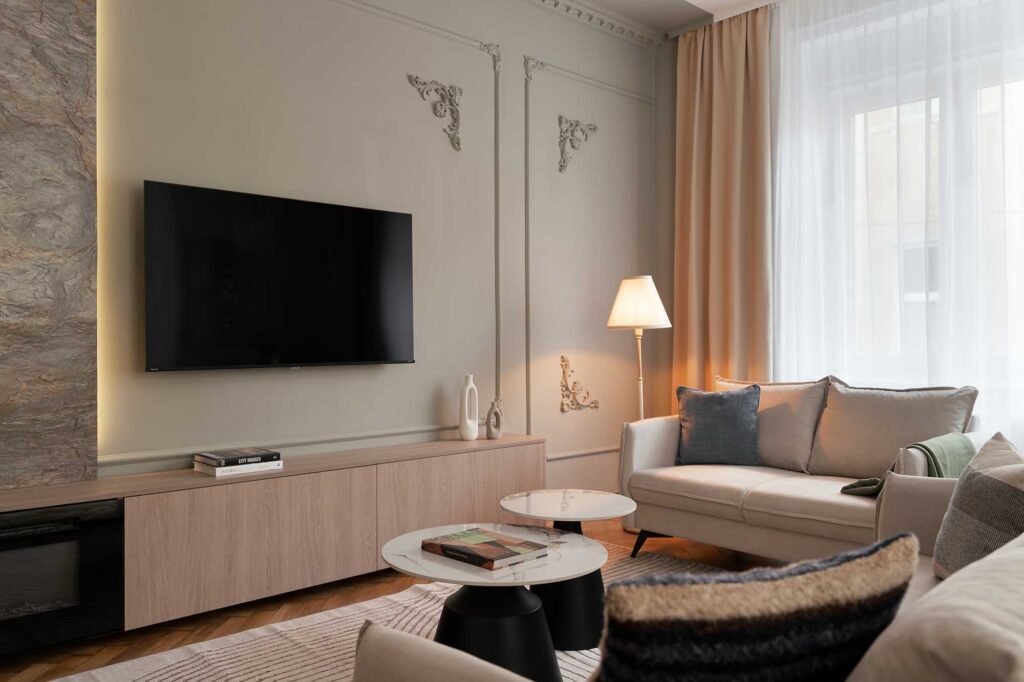 Elegant modern living room with beige sofa, marble coffee tables, and ornate wall moldings featuring a textured stone panel and mounted TV, complemented by soft curtains, warm floor lamp, and natural light enhancing the neutral color palette.