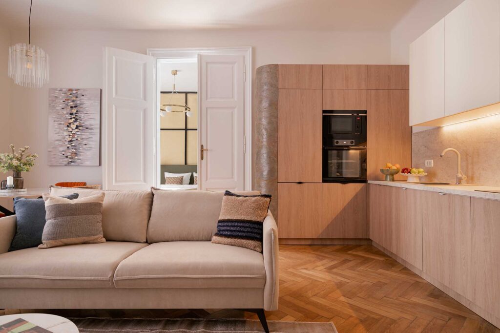 Contemporary open‑plan living space captured by interior photographer ilisie sergiu, with beige sofa, textured cushions, and light wood kitchen cabinetry featuring built‑in oven, marble backsplash, and warm under‑cabinet lighting, complemented by herringbone parquet flooring and natural light through tall windows.