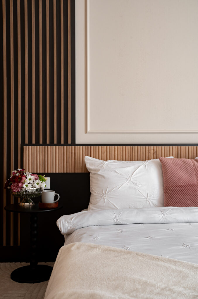 Editorial hospitality photograph capturing a boutique bedroom with soft white bedding, blush accent pillow, and vertical wood paneling, styled to evoke quiet luxury and intimate comfort — ideal for hotels and guesthouses seeking refined visual storytelling