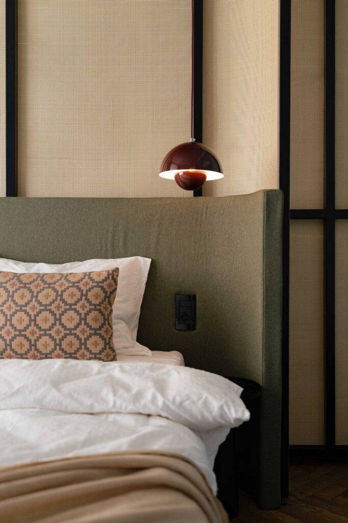 Close‑up editorial photo captured by Ilisie Sergiu of a modern bedroom with green upholstered headboard, geometric wall panels in beige and black, and a brown dome pendant light, featuring a decorative pillow with woven geometric texture in beige and terracotta tones, white bedding, and soft beige throw.