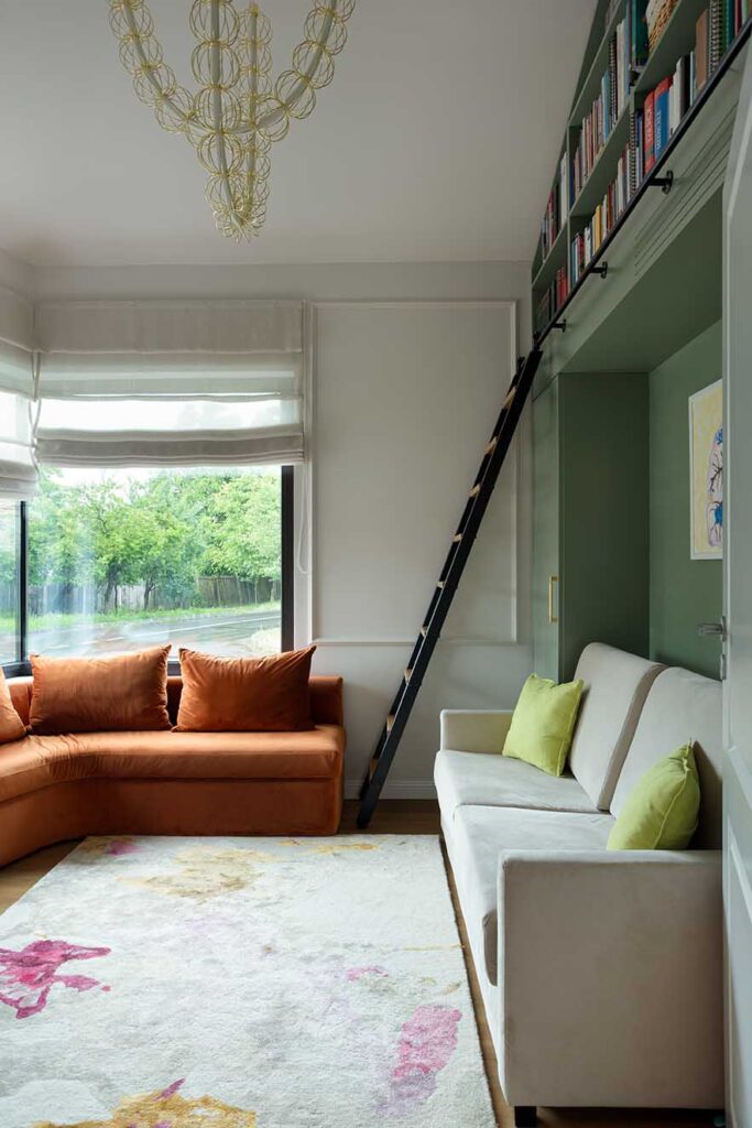 "Contemporary reading lounge featuring a burnt‑orange sectional and cream sofa beneath a built‑in bookshelf with ladder access. A balanced composition that attracts interior designers through its layered color palette, architectural integration, and inviting natural light.