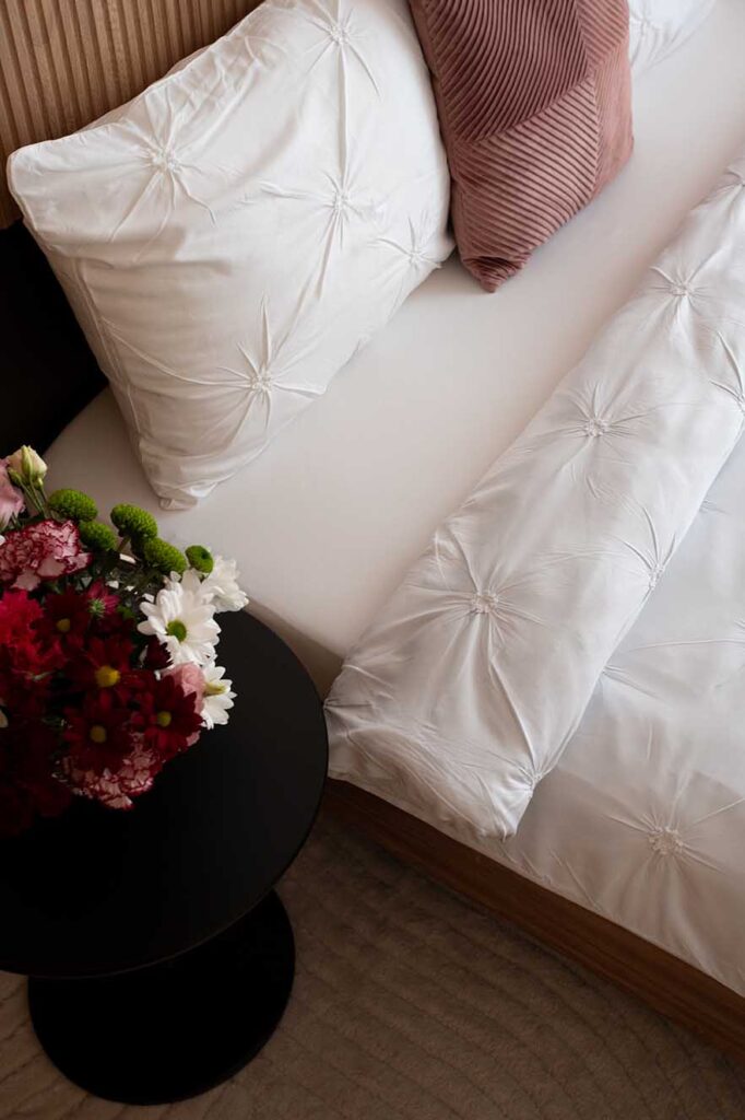 Hospitality Photography Close‑up of modern bedroom with white gathered bedding, ribbed rose pillow, and round black bedside table holding a vibrant floral arrangement of red, white, and green blooms, complemented by a beige ridged headboard and soft neutral tones.