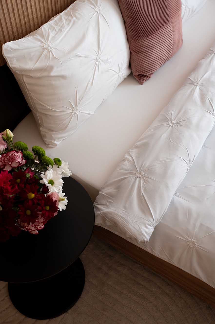 Hospitality Photography Close‑up of modern bedroom with white gathered bedding, ribbed rose pillow, and round black bedside table holding a vibrant floral arrangement of red, white, and green blooms, complemented by a beige ridged headboard and soft neutral tones.