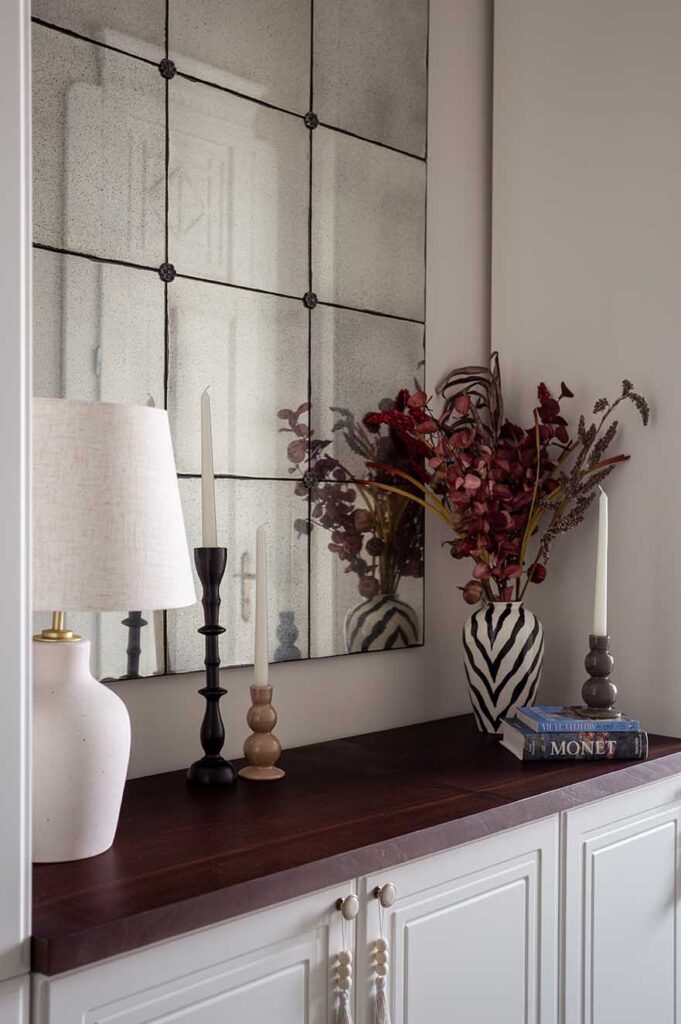 Curated console vignette with an aged paneled mirror, sculptural ceramic lamp, striped monochrome vase with dried foliage, and art books including a Monet volume. A balanced study in texture, proportion, and refined decorative layering.