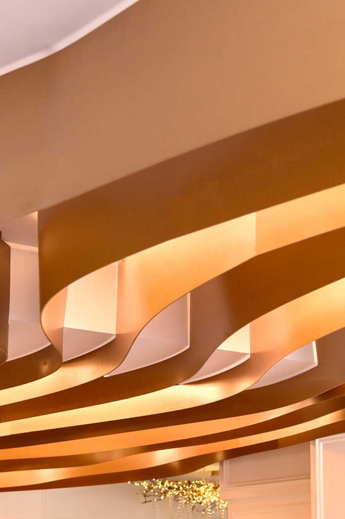 Curved ceiling installation in warm bronze tones with integrated lighting, showcasing sculptural rhythm and soft illumination. A statement detail appealing to interior designers focused on expressive architectural forms.